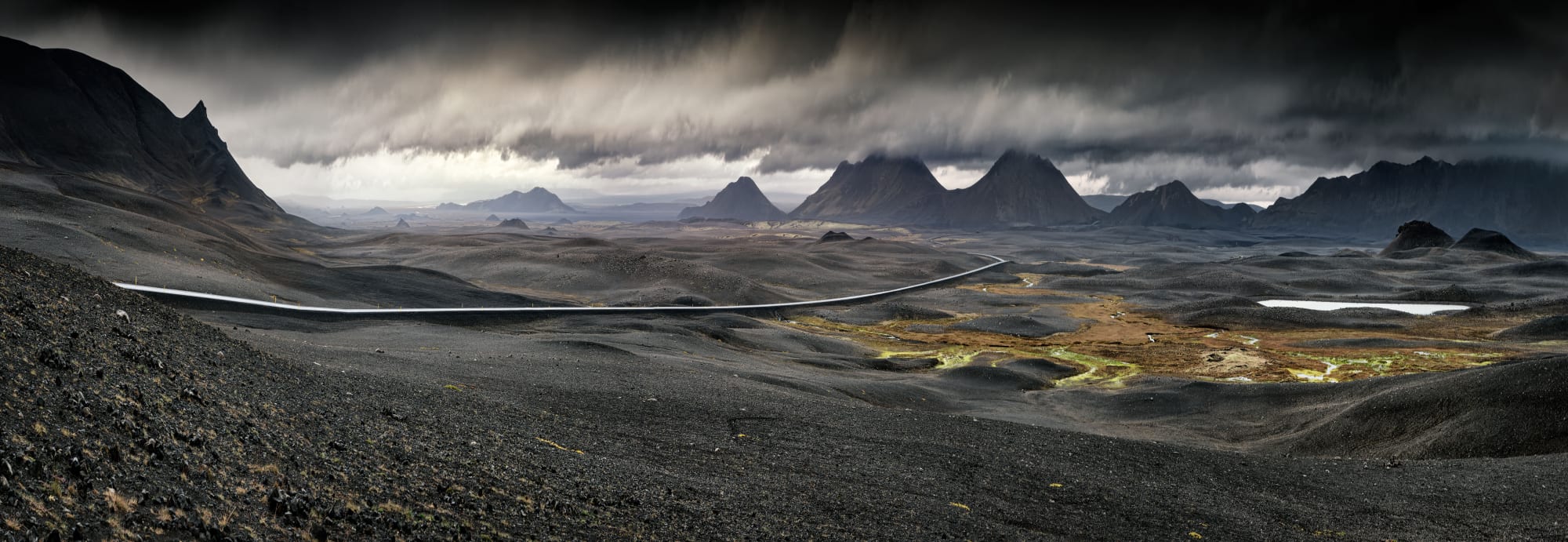 A sweeping panoramic view of Iceland's stark volcanic highlands, featuring a winding glacial river cutting through dark lava fields, dramatic jagged mountain peaks in the distance, and moody storm clouds overhead. The landscape showcases the raw, otherworldly terrain characteristic of Iceland's interior, with patches of golden vegetation dotting the desolate terrain.