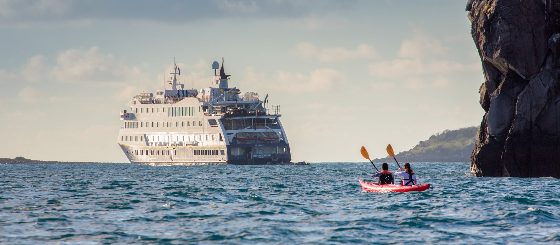 Two kayakers in a red kayak paddle near dramatic limestone cliffs while a large white cruise ship