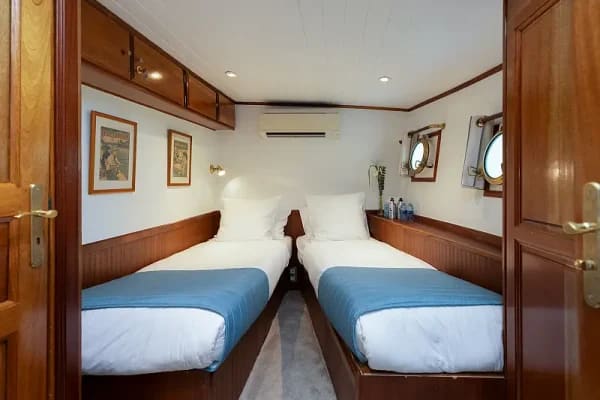 A compact twin-bed cabin aboard a cruise ship featuring two single beds with blue covers, wooden cabinetry, white walls, and nautical-style portholes. The space exemplifies efficient cruise ship accommodation design with warm wood tones and practical furnishings typical of mid-range cruise vessel interiors.