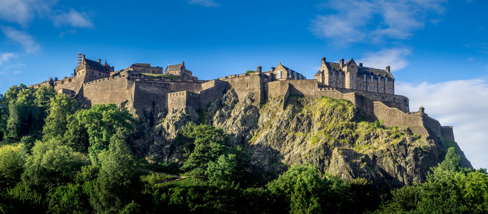 Edinburgh Castle stands majestically atop a dramatic volcanic crag overlooking the city of Edinburgh, Scotland. The historic fortress features distinctive stone architecture with turrets and defensive walls, surrounded by lush green vegetation on the steep rocky outcrop.