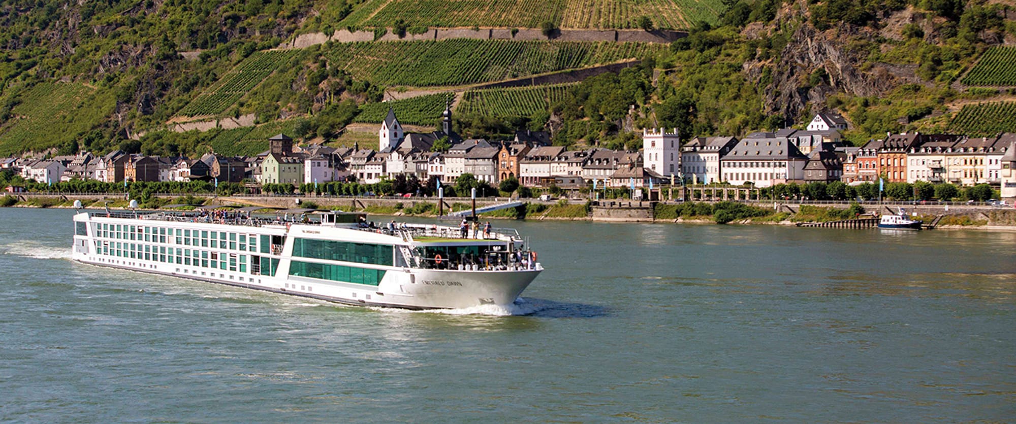 A white and green river cruise ship sailing past a historic European town with vineyards