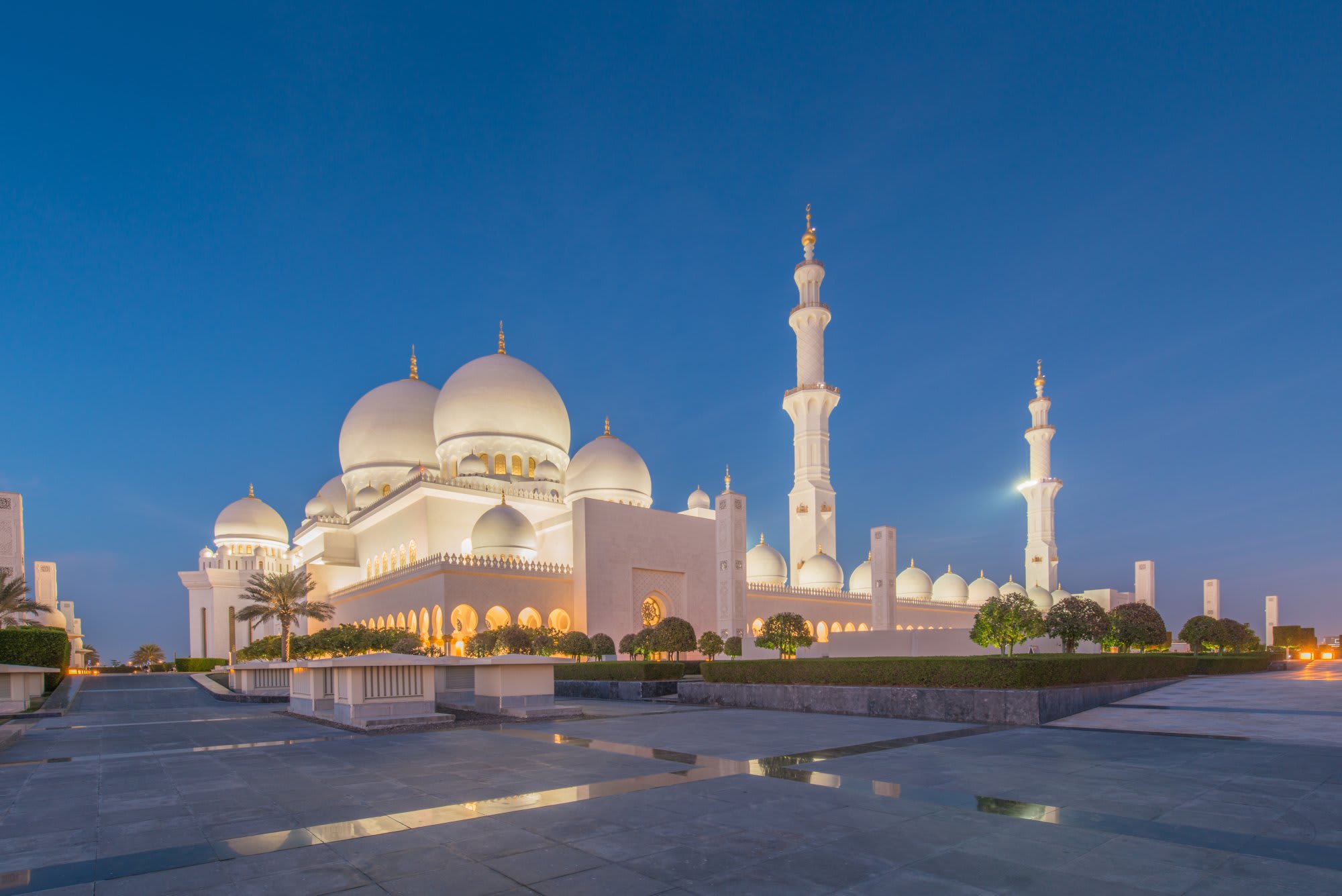 The iconic Sheikh Zayed Grand Mosque illuminated at dusk against a deep blue sky, featuring pristine white marble architecture with multiple domes and minarets, reflected in the still water of the courtyard plaza. The mosque's stunning Islamic design and symmetrical layout showcase the architectural grandeur of Abu Dhabi's most significant religious landmark.