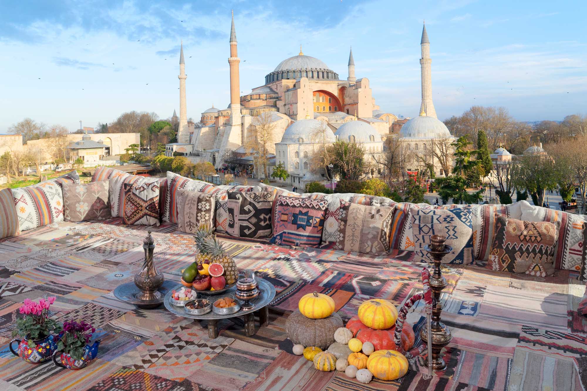 A stunning rooftop terrace featuring traditional Turkish cushions, kilim textiles, and autumn produce overlooking the iconic Hagia Sophia mosque in Istanbul, Turkey. The scene captures the architectural grandeur of the historic landmark with its massive dome and minarets against a clear blue sky.