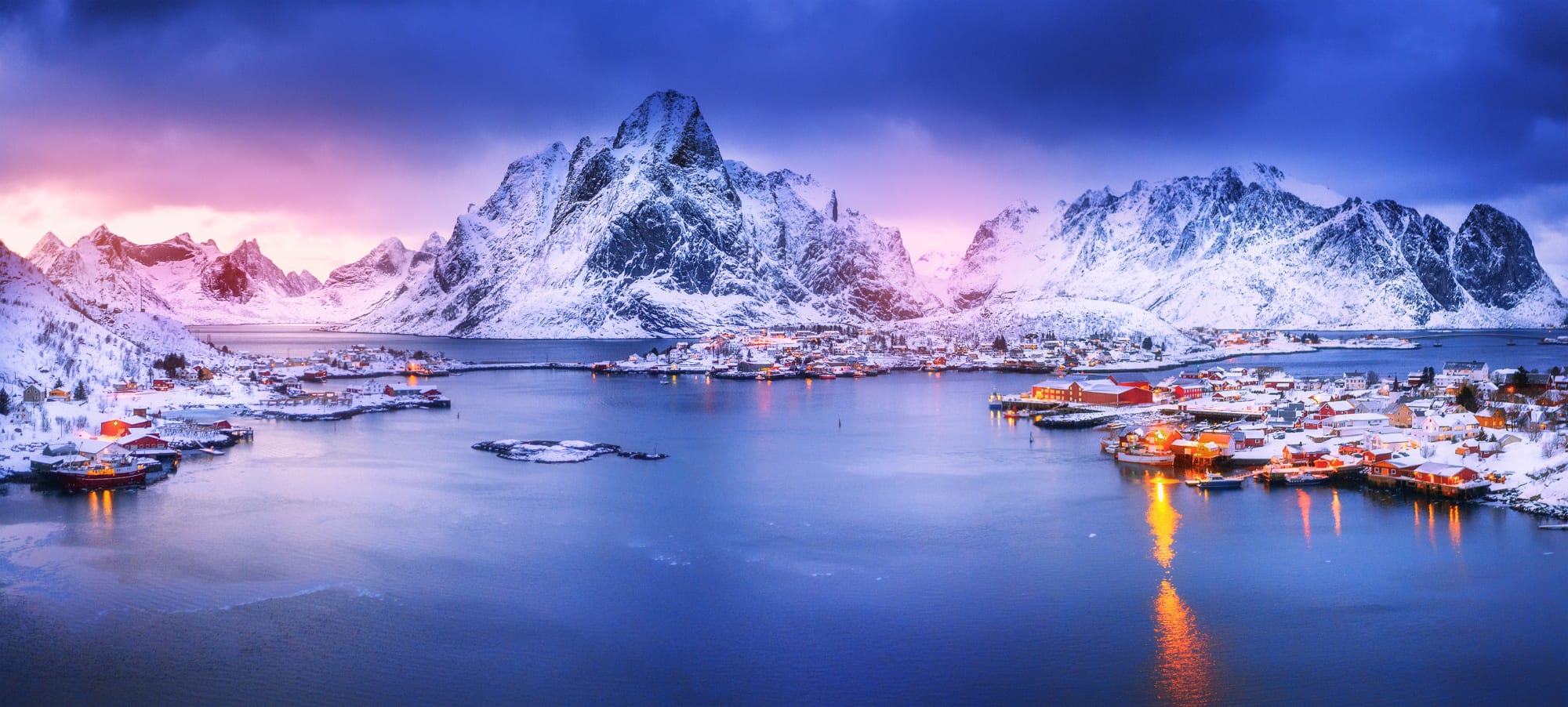 A stunning winter landscape featuring snow-capped mountains towering over a picturesque Arctic fishing village with traditional red and yellow wooden houses reflected in calm blue waters during twilight. The scene captures the dramatic beauty of Norway's Lofoten Islands with their characteristic peaks and traditional fishermen's cabins (rorbuer) illuminated by the soft glow of sunset.