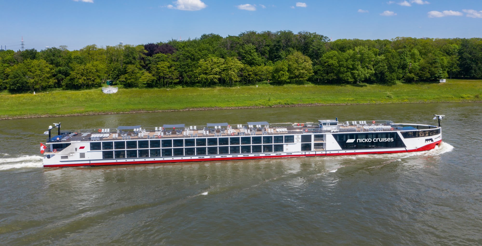 A modern Nicko Cruises river cruise ship navigates a European river with lush green countryside visible along the banks. The vessel features extensive deck cargo and continuous window lines characteristic of contemporary river cruise design.