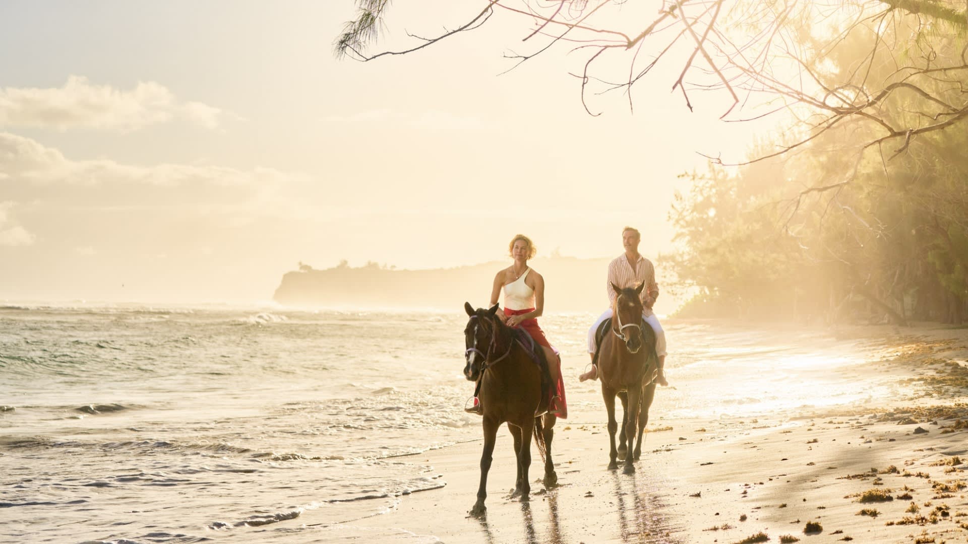 Two riders on horseback traverse a pristine sandy beach at golden hour, with gentle waves lapping