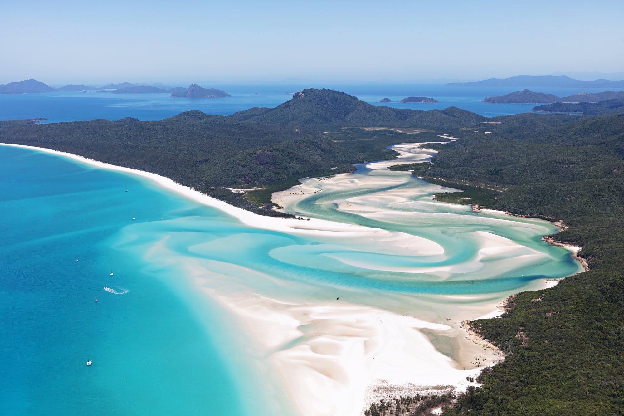 An aerial panorama of Whitehaven Beach on Whitsunday Island, Australia, showcasing the iconic white silica sand beach with turquoise lagoons, surrounding rainforest-covered hills, and numerous small islands dotting the sapphire waters of the Coral Sea.