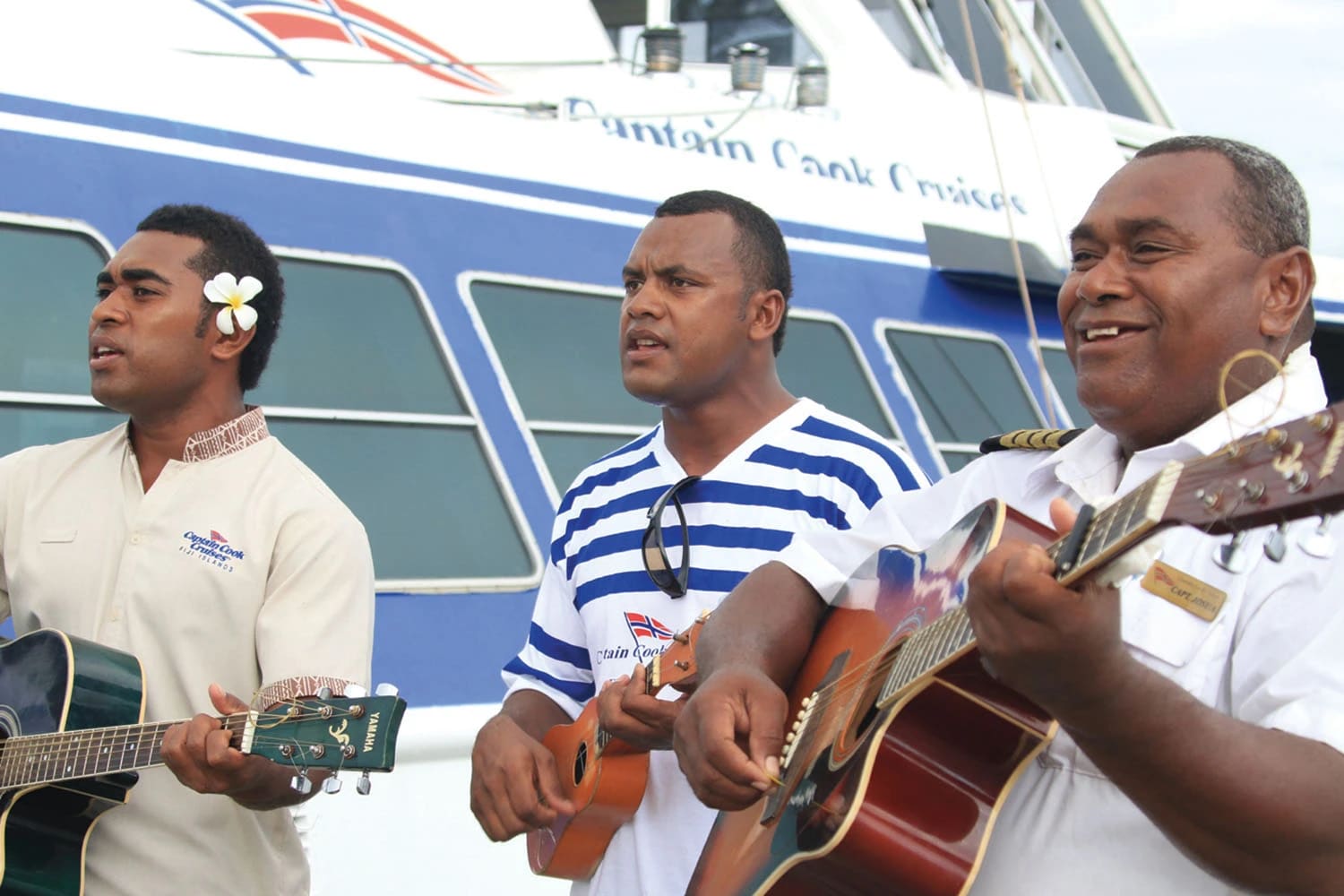 Three musicians perform together with acoustic guitars on the deck of a Dawain Bank Cruises vessel