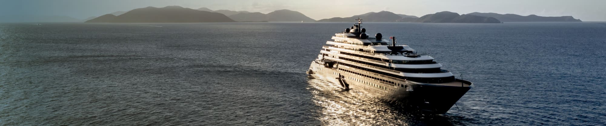 A sleek, multi-deck white luxury yacht sailing on calm blue waters with a mountainous coastline in the background. The vessel appears to be a high-end private yacht with multiple elegant curved decks.