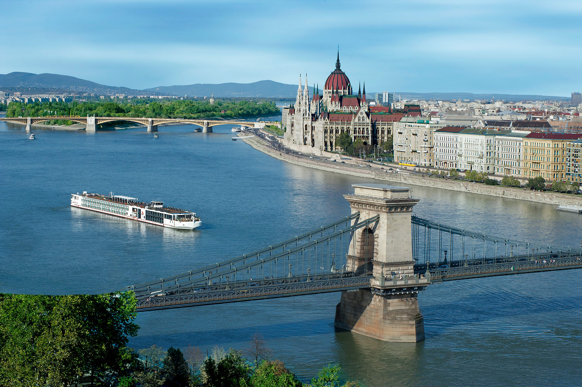 A modern river cruise vessel is moored along a tree-lined riverbank under a partly cloudy sky
