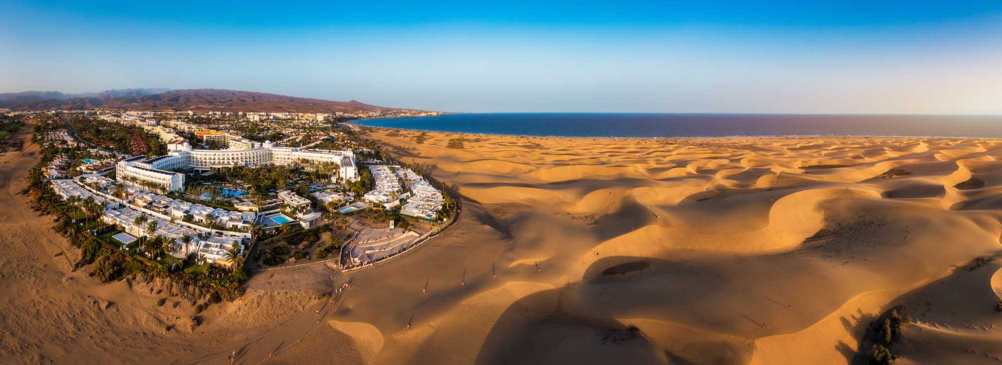 An aerial view of the golden sand dunes of Maspalomas Natural Reserve in Gran Canaria, with a luxurious beachfront resort nestled on the coast overlooking the Atlantic Ocean and surrounding desert landscape.