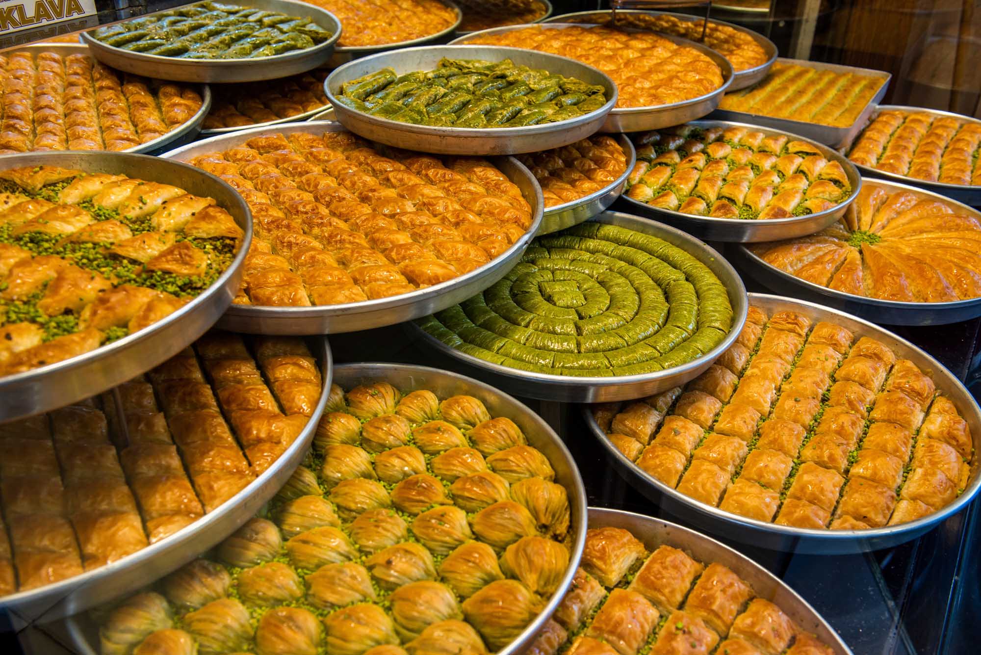 A vibrant display of various traditional Turkish baklava varieties arranged in metal trays at a dessert shop. The golden pastries are garnished with pistachios and arranged in decorative patterns, showcasing different baklava styles including spiral-shaped pistachio baklava and rectangular honey-soaked varieties typical of Gaziantep, Turkey.