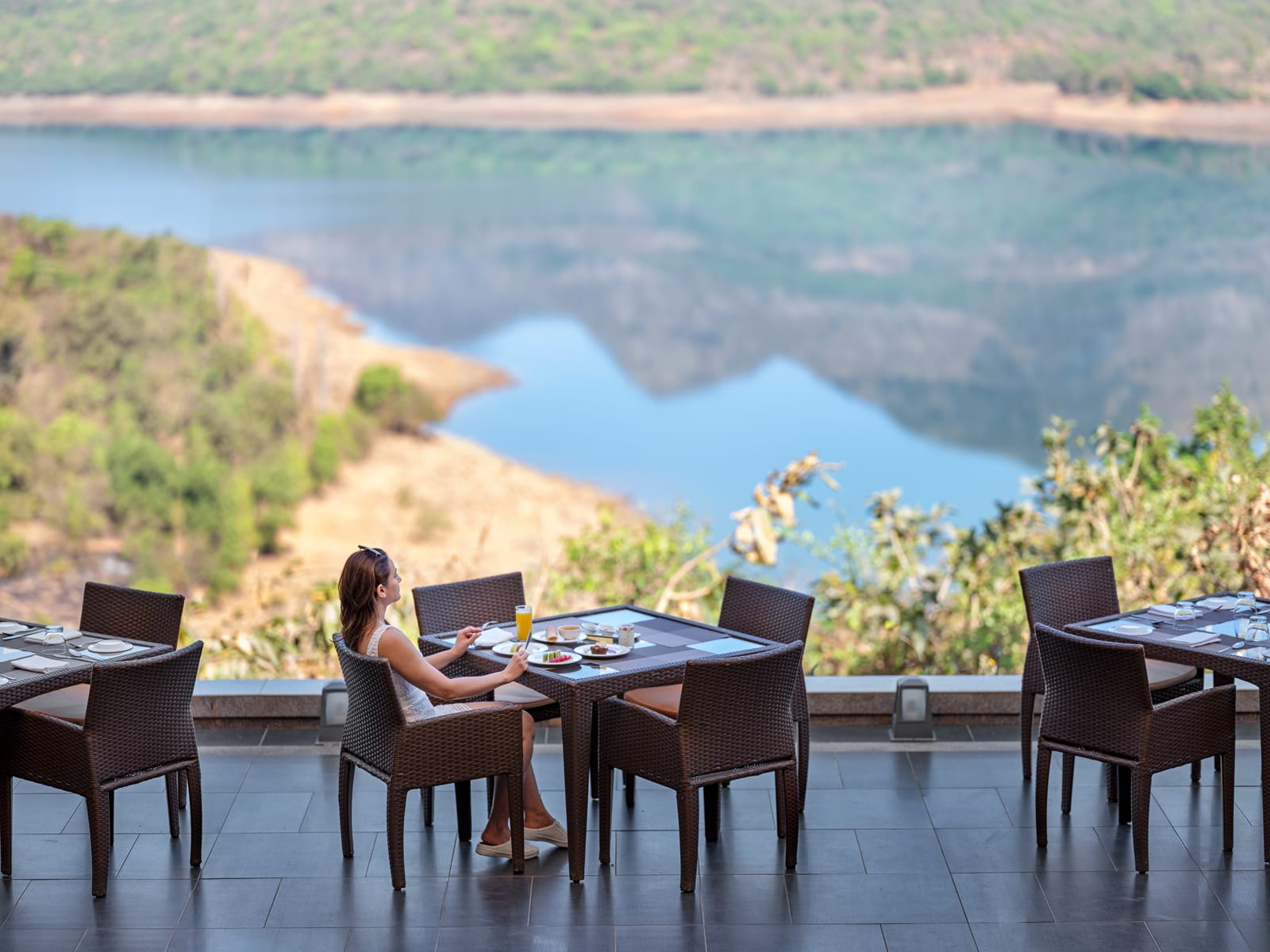 Woman dining alone at outdoor table overlooking turquoise lake and forested hillside