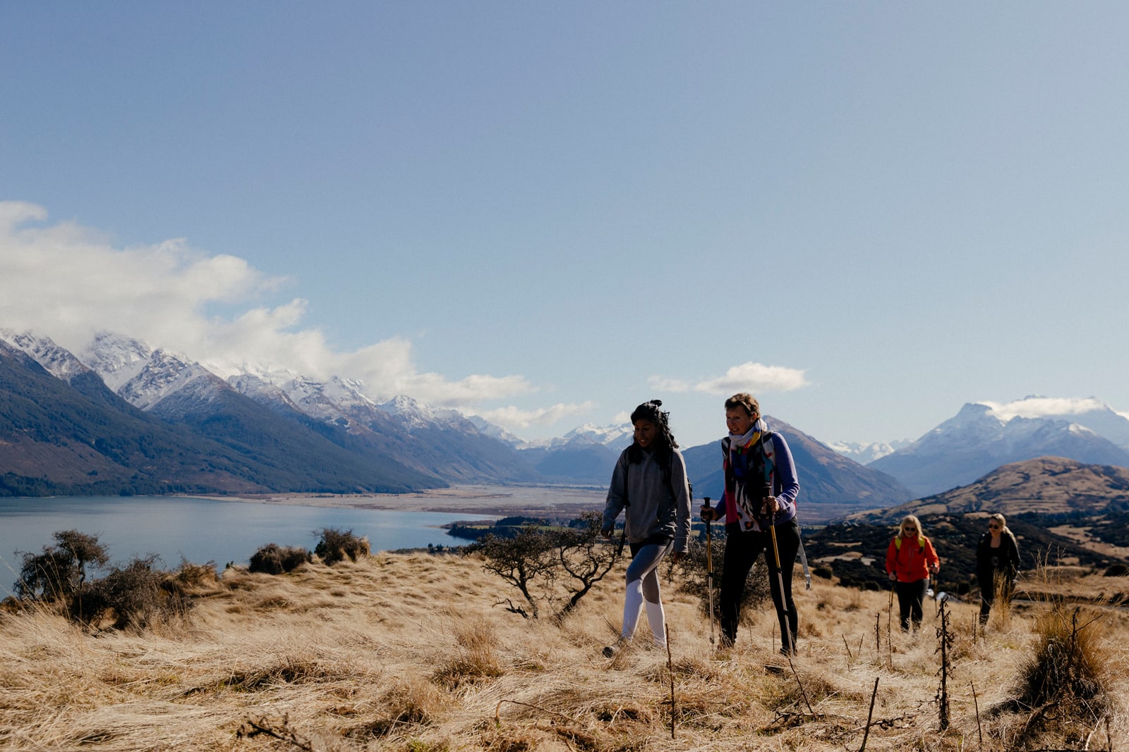 Group of hikers on dry hillside with snow-capped mountains and lake in background