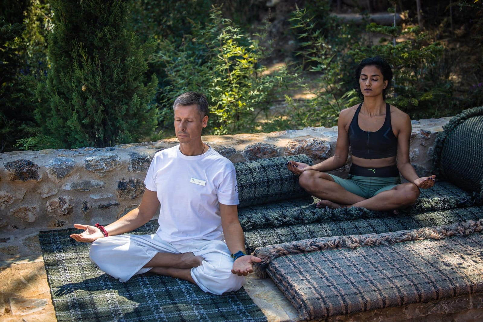 Two people meditating outdoors on woven mats among lush green garden surroundings