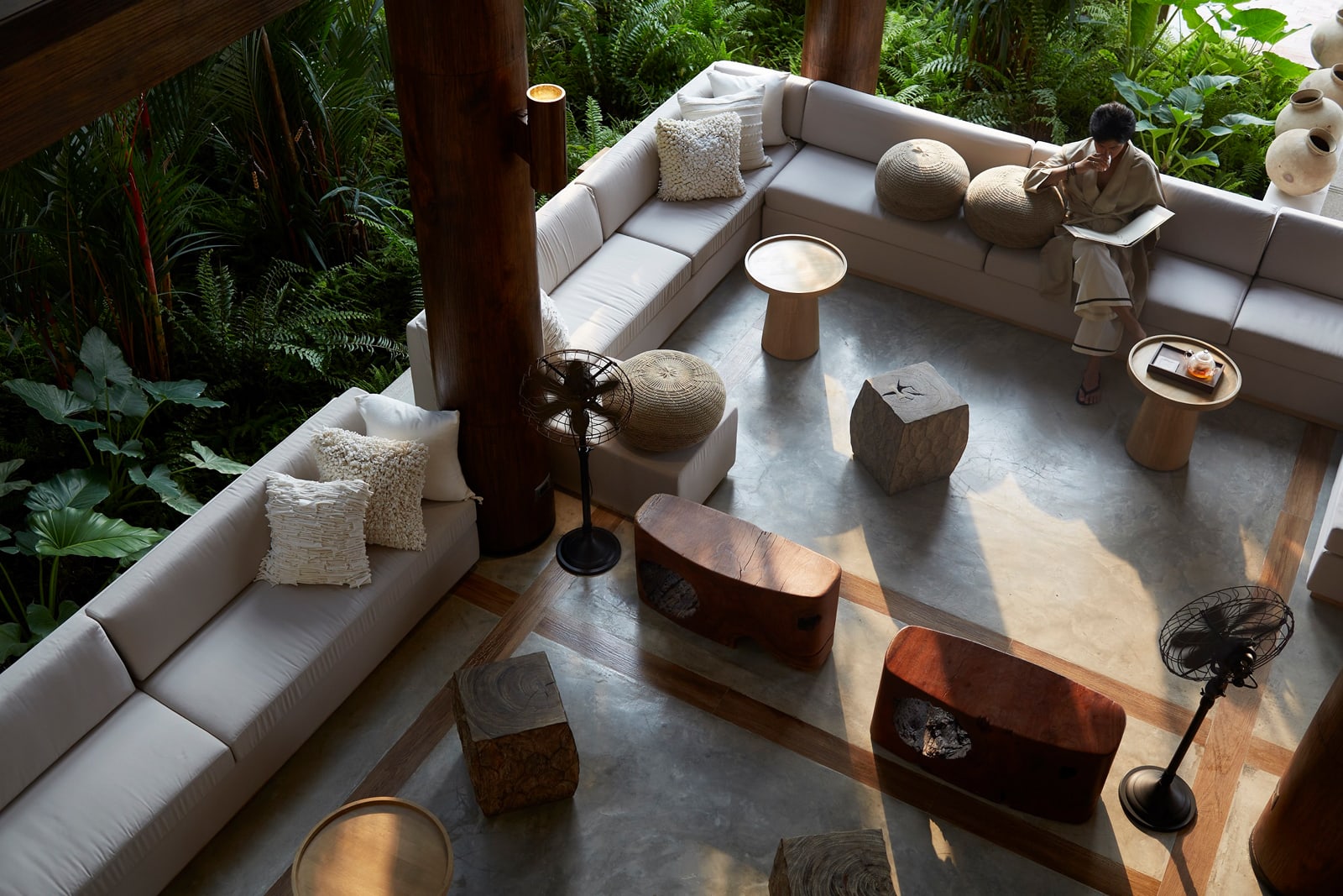 A contemporary interior living space with minimalist white sofas, wooden accents, and floor-to-ceiling windows overlooking a dense tropical forest environment. The space features organic design elements and natural materials, creating a serene indoor-outdoor connection.