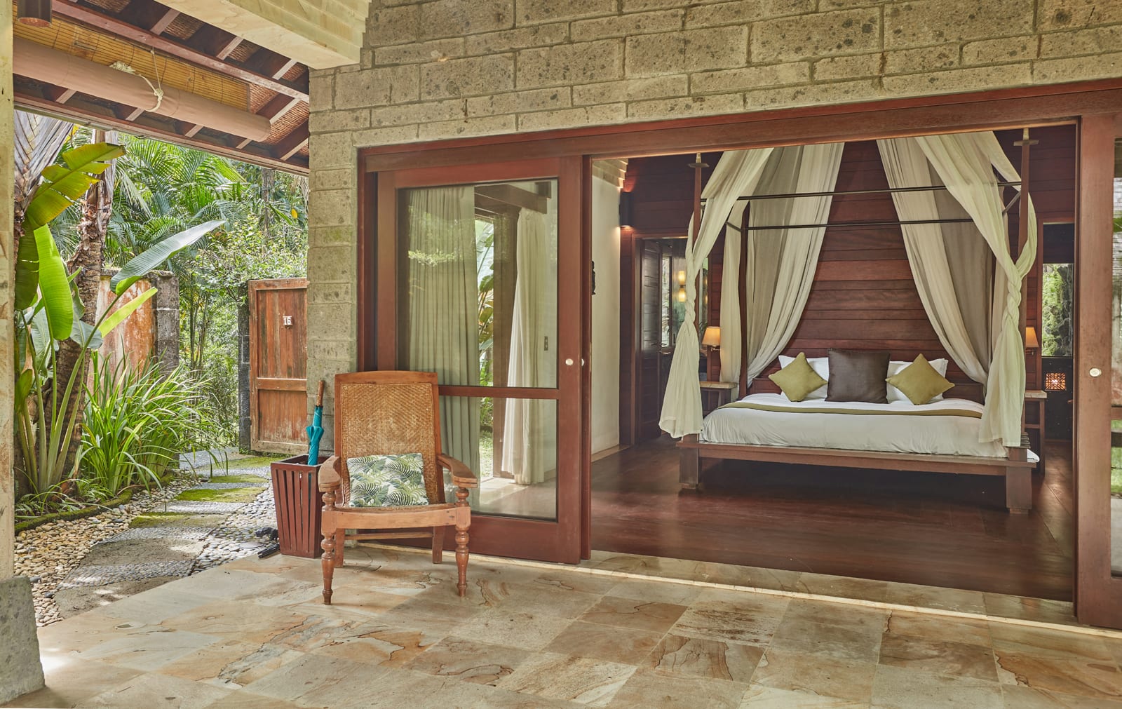 Luxury tropical bedroom with wooden four-poster bed, cream curtains, and garden view through open glass doors