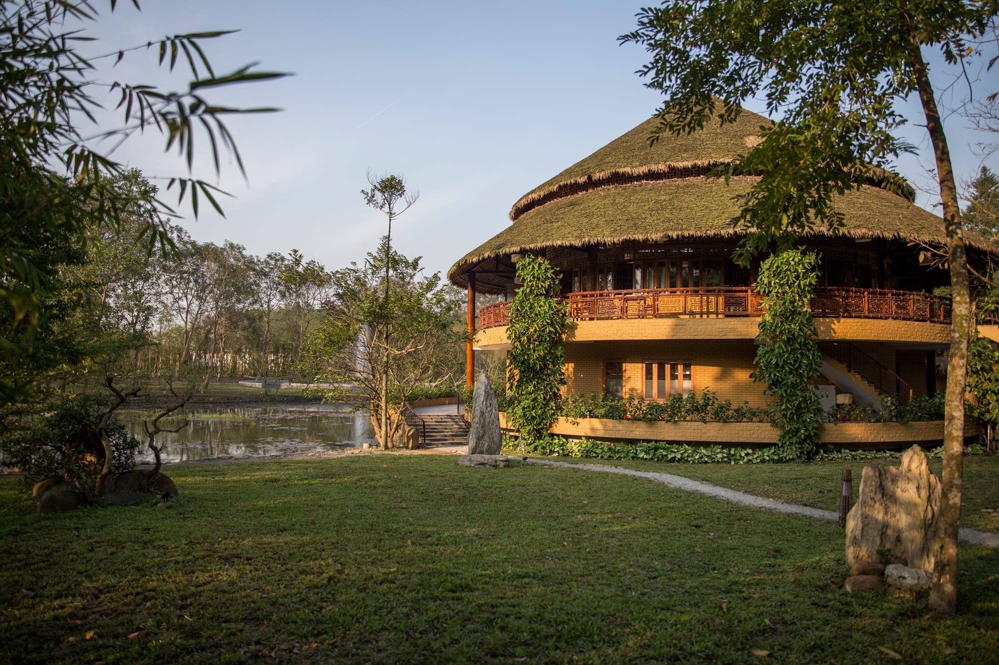 Round thatched-roof lodge with wooden railings overlooking a pond surrounded by tropical trees