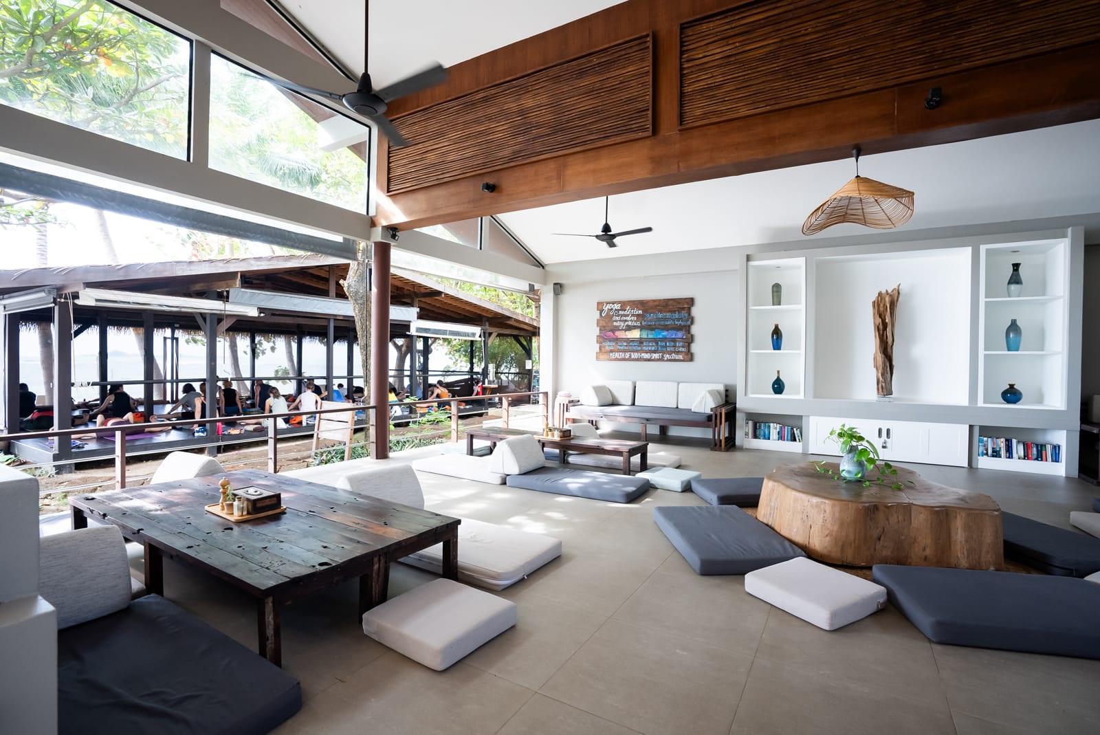 Modern open-plan lounge with wooden beams, floor cushions, and outdoor seating area visible through glass doors