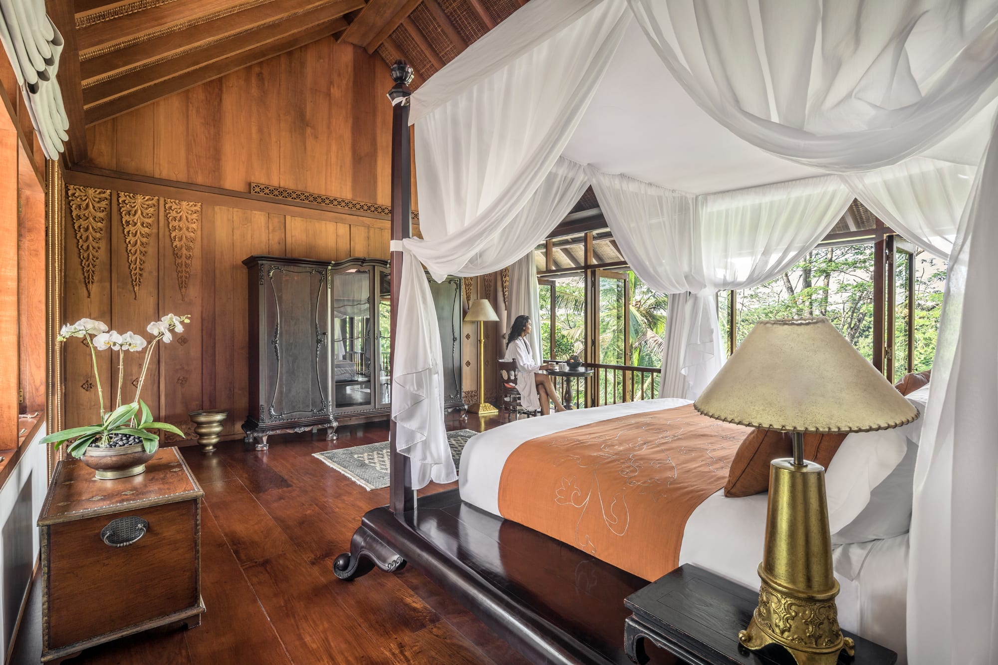 Luxury bedroom with four-poster canopy bed, white sheer curtains, dark wood furniture, and forest views through large windows