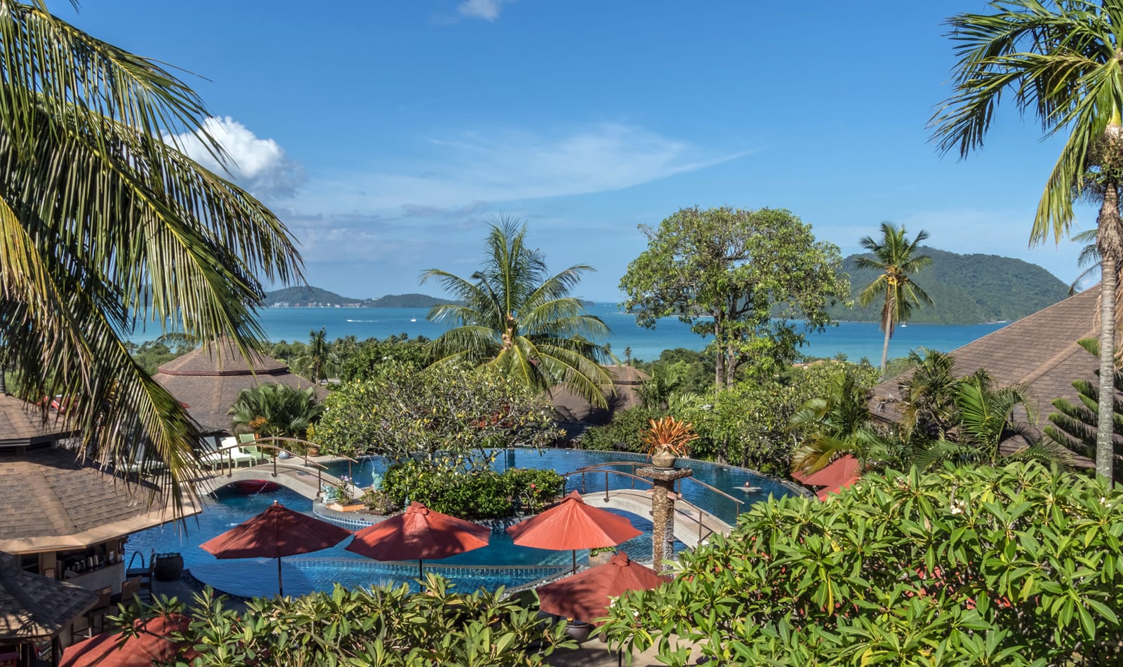 Tropical resort pool with red umbrellas overlooking turquoise bay and palm-forested coastline
