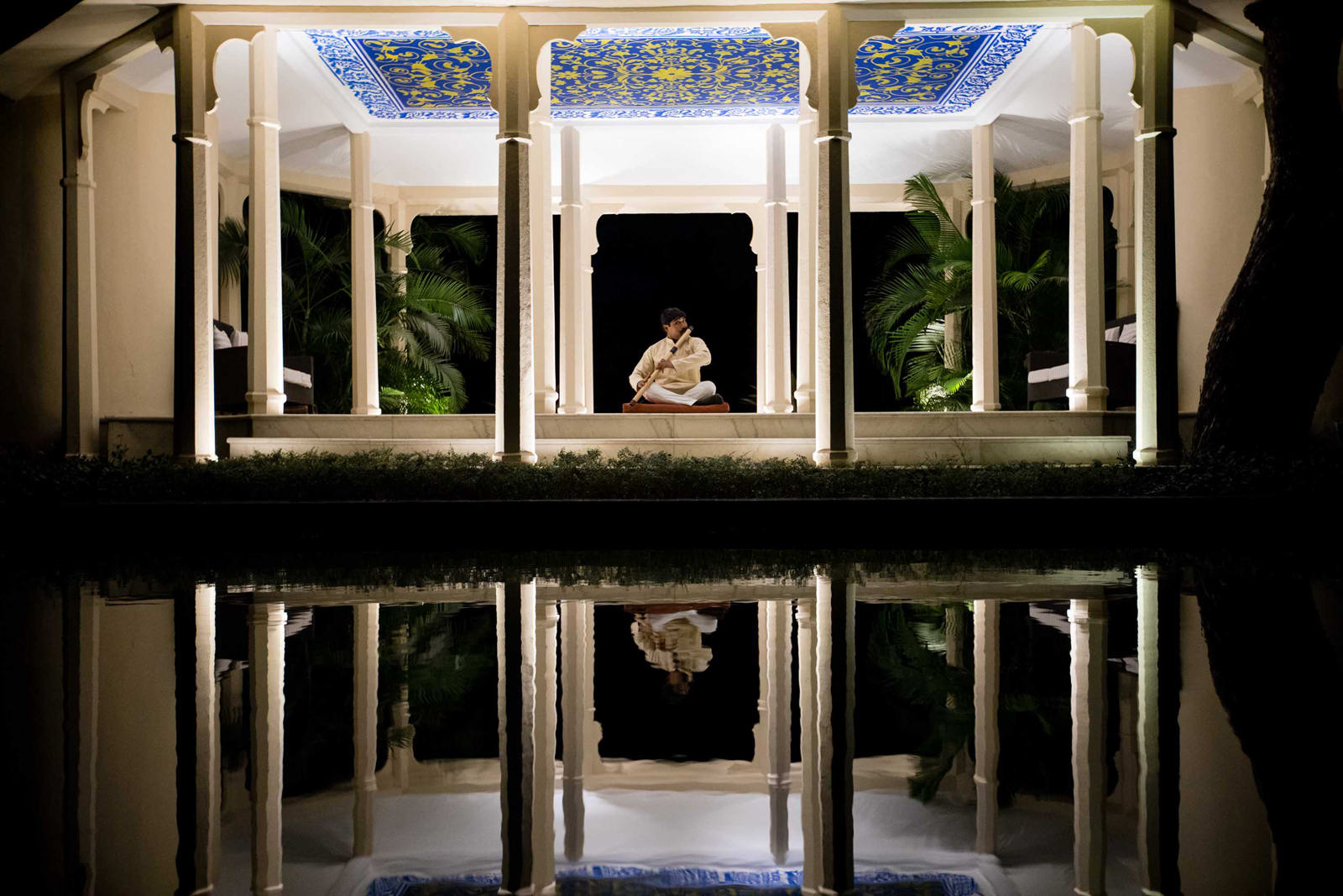 An elegant architectural space with white columns and ornate blue-and-yellow ceiling panels, featuring a figure seated in meditation with lush greenery and a perfect water reflection.