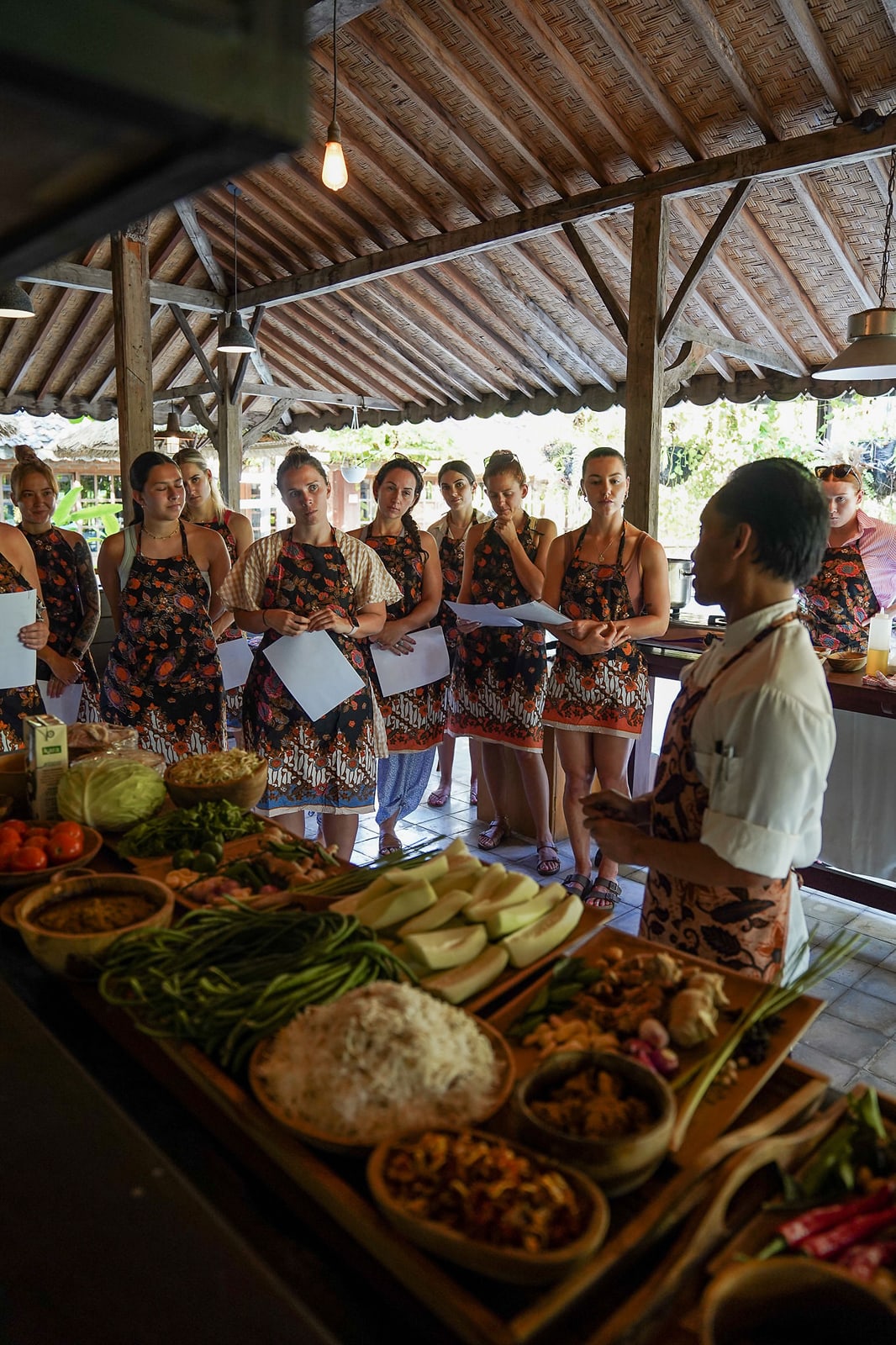 Group of women in matching batik aprons learning traditional cooking in covered pavilion with fresh ingredients displayed
