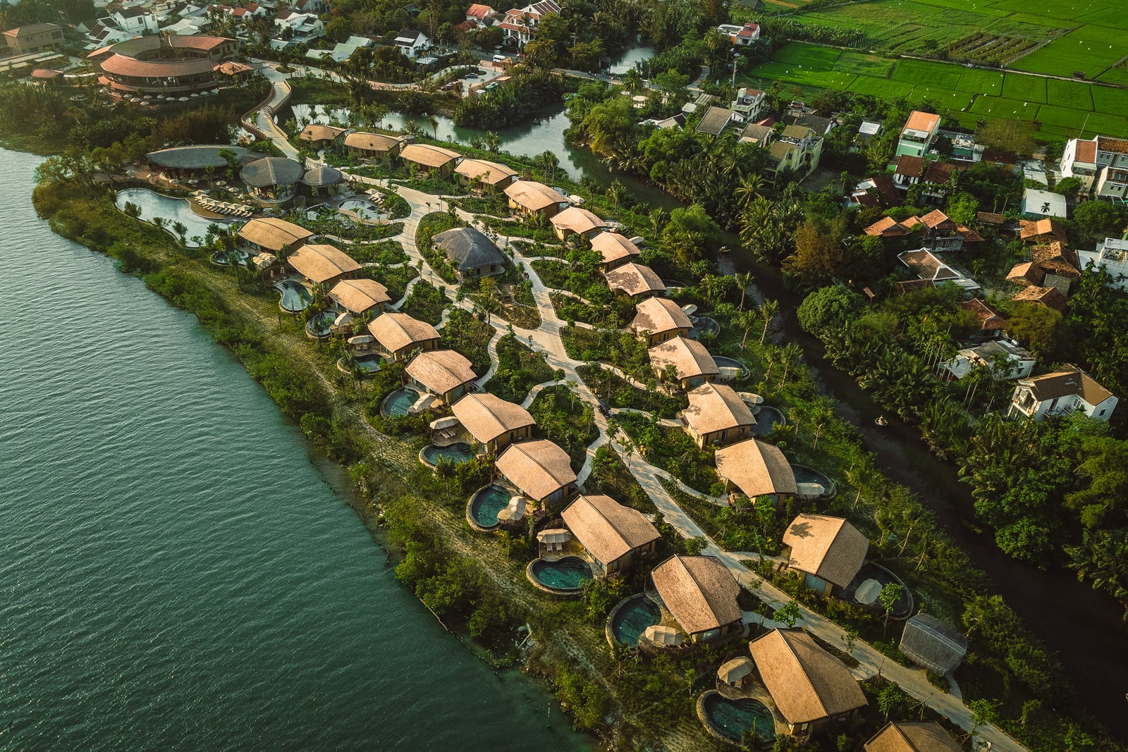 Aerial view of riverside resort with thatched-roof bungalows and private pools along turquoise water