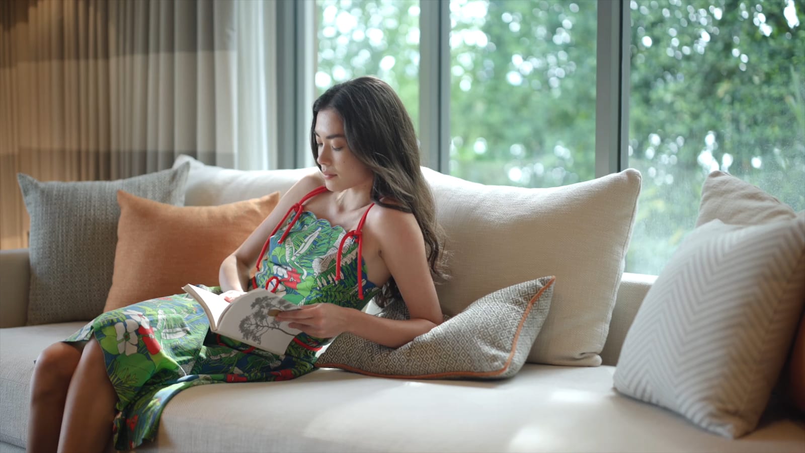 Woman in tropical print dress reading book on beige sofa by large windows with garden view