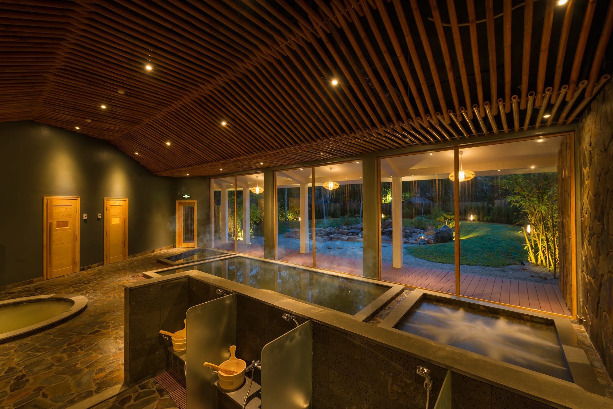 Modern indoor onsen spa with wooden lattice ceiling, stone soaking tubs, and garden views at night