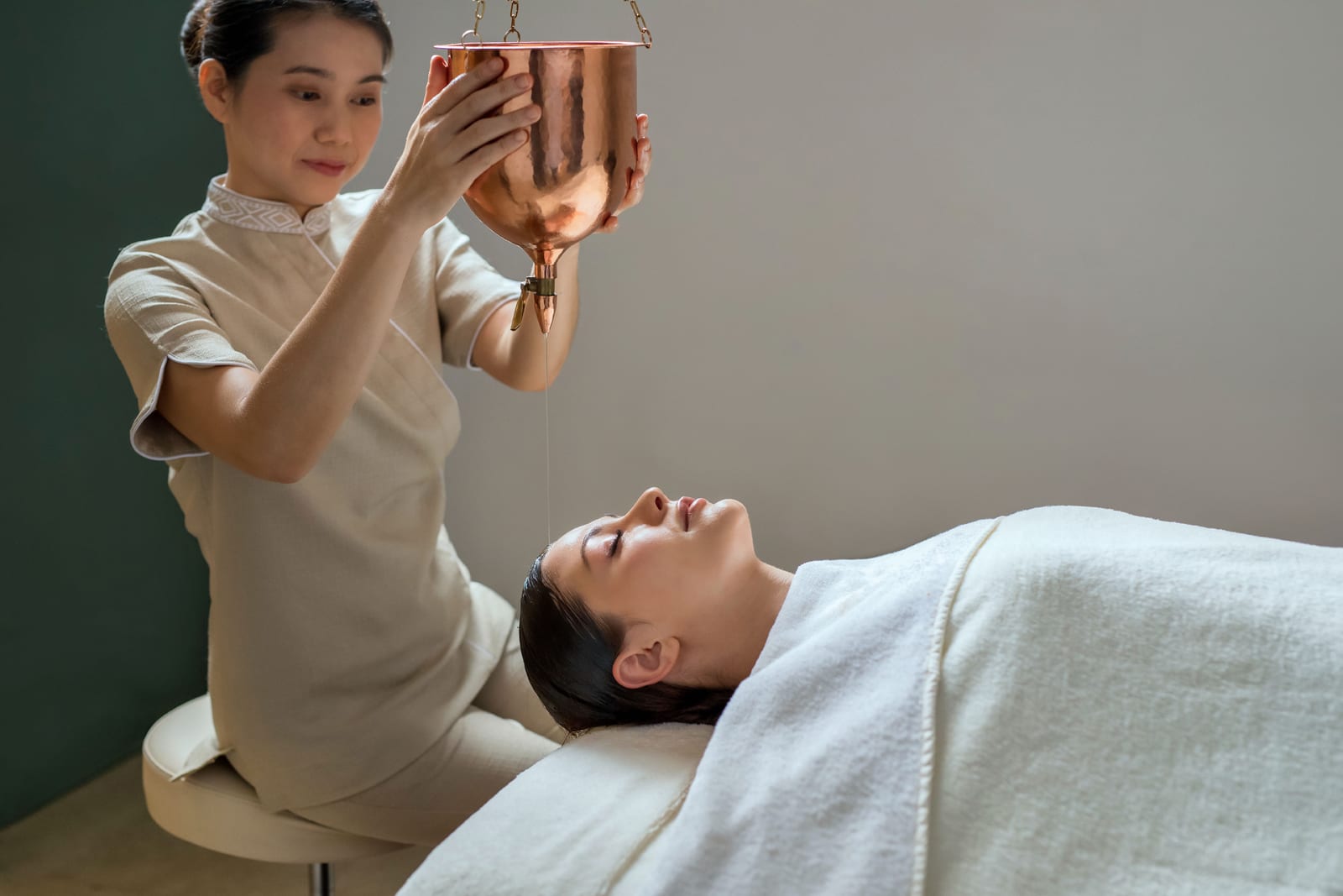 Therapist performing shirodhara, pouring warm oil on reclined client's forehead