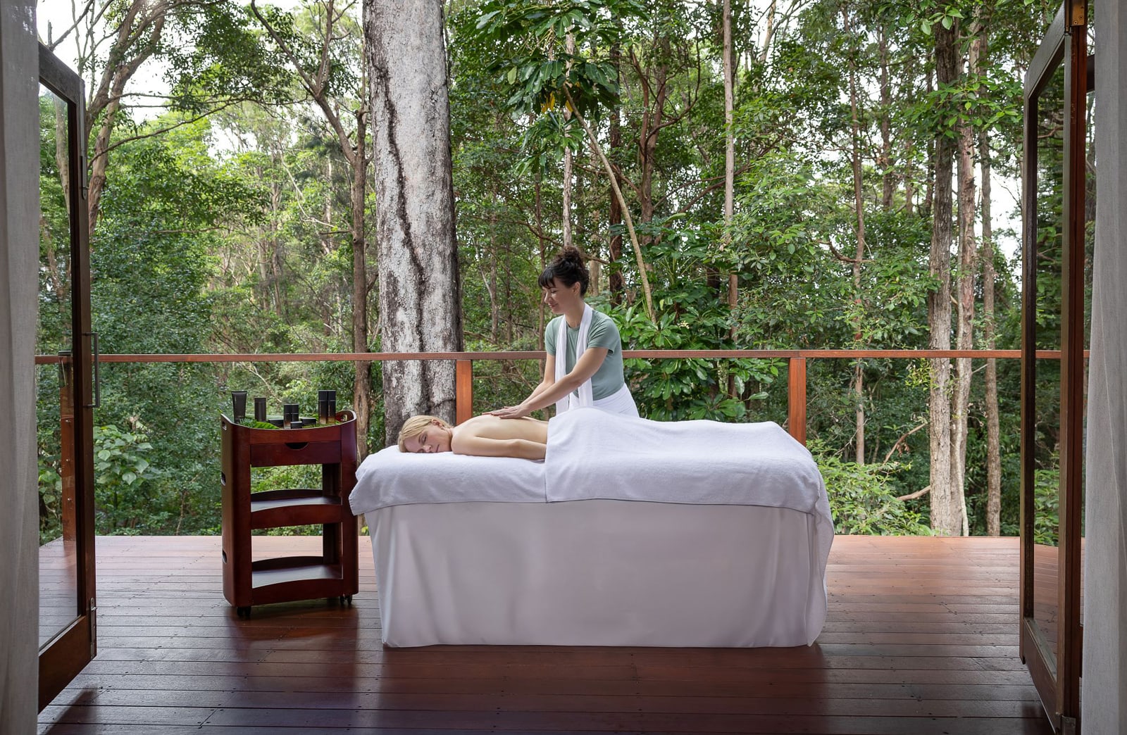 Therapist performing back massage on client in open-air spa pavilion surrounded by forest trees