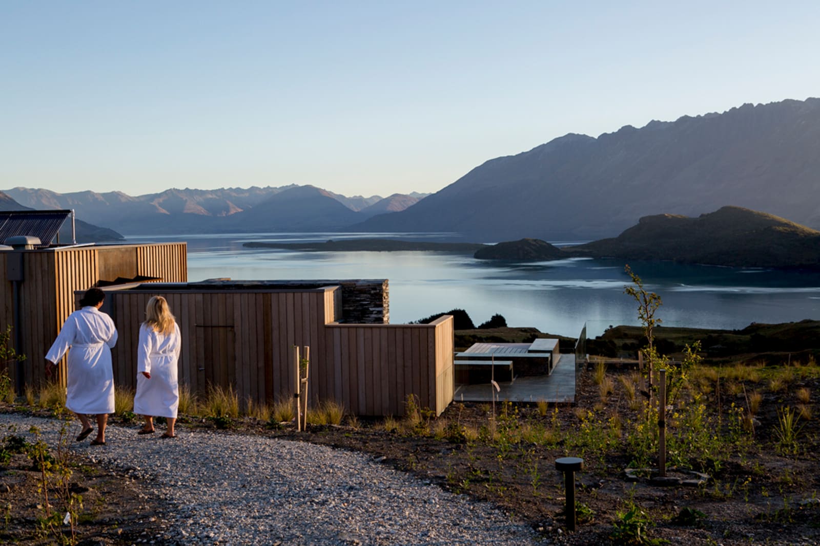 Two people in white robes walking toward modern cabin with mountain lake view