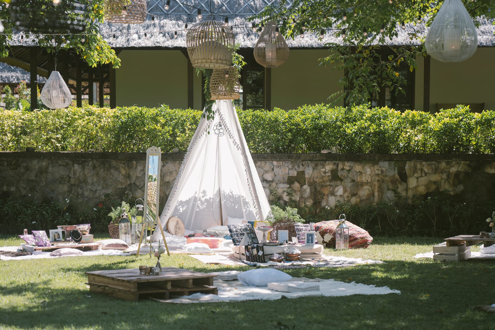 Bohemian garden picnic setup with white teepee tent, woven hanging lanterns, and scattered cushions on grass surrounded by hedges