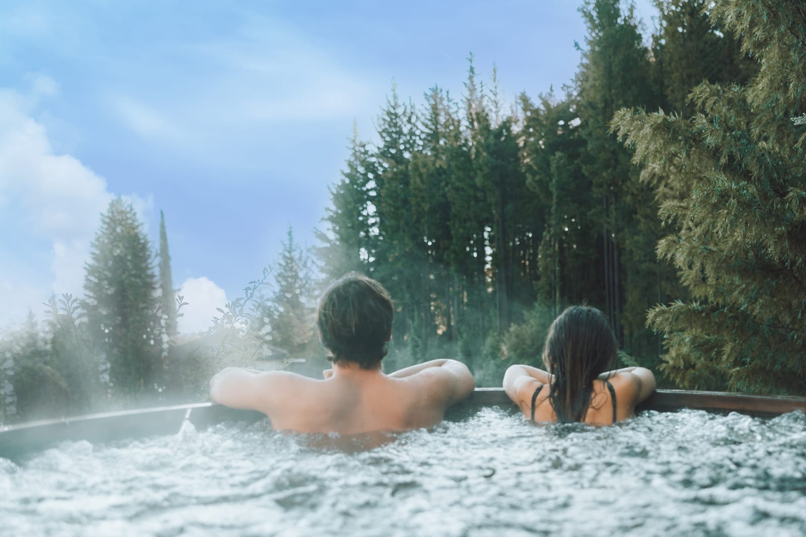 Two people relaxing in steaming outdoor hot tub surrounded by tall evergreen trees