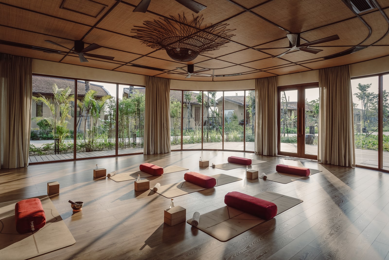 Yoga studio with burgundy bolsters on mats, woven ceiling fixture, and floor-to-ceiling windows overlooking tropical gardens