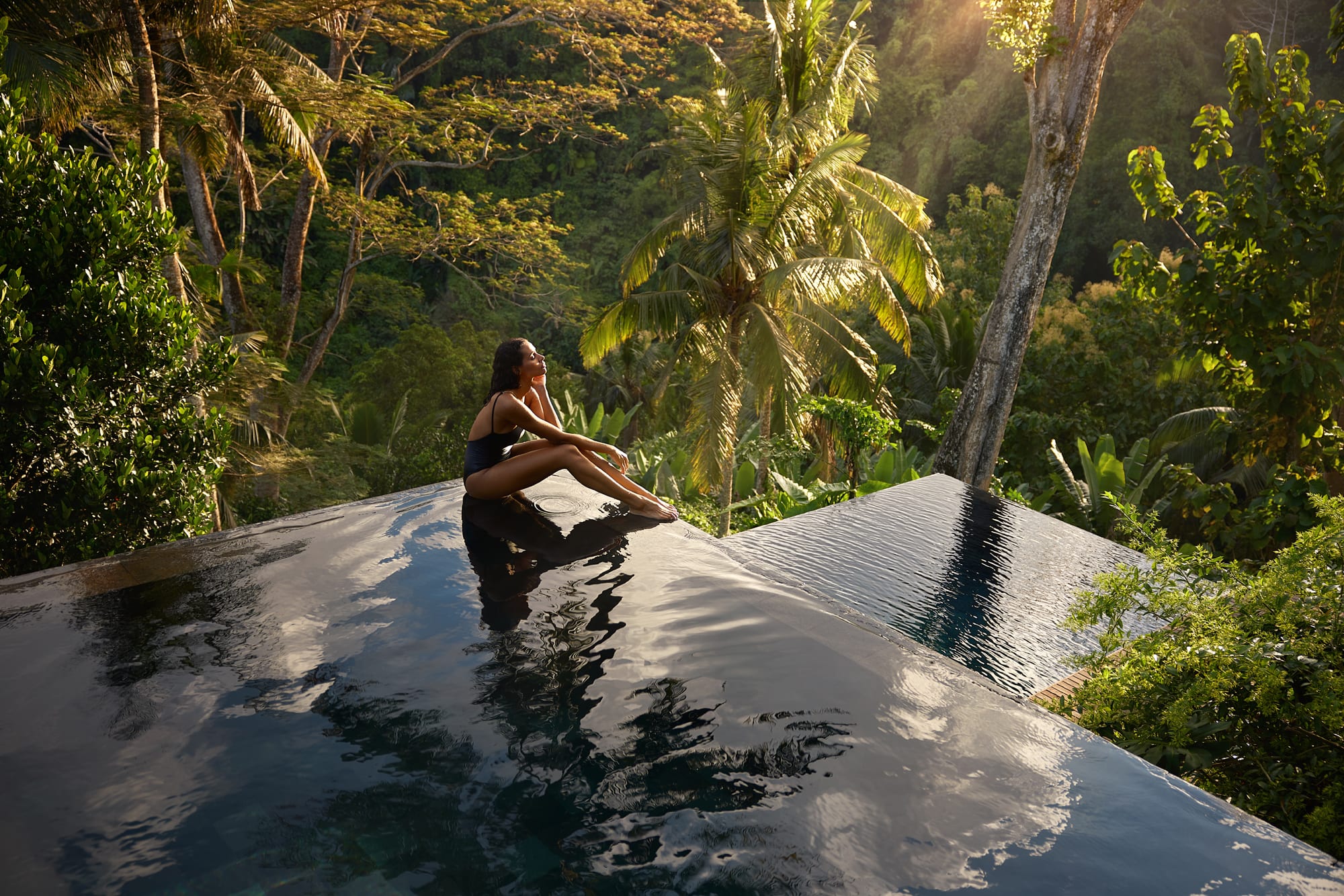 Woman relaxing in infinity pool overlooking lush tropical forest valley at sunrise
