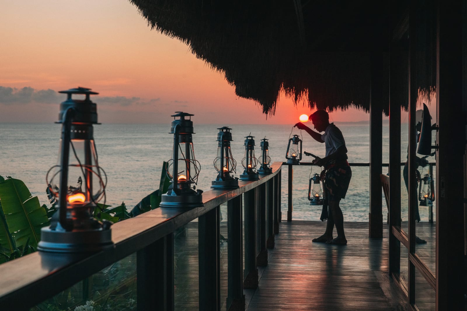 A serene scene of vintage lanterns being lit on a wooden pier at sunset, with a thatched roof overhead and the ocean stretching to the horizon.