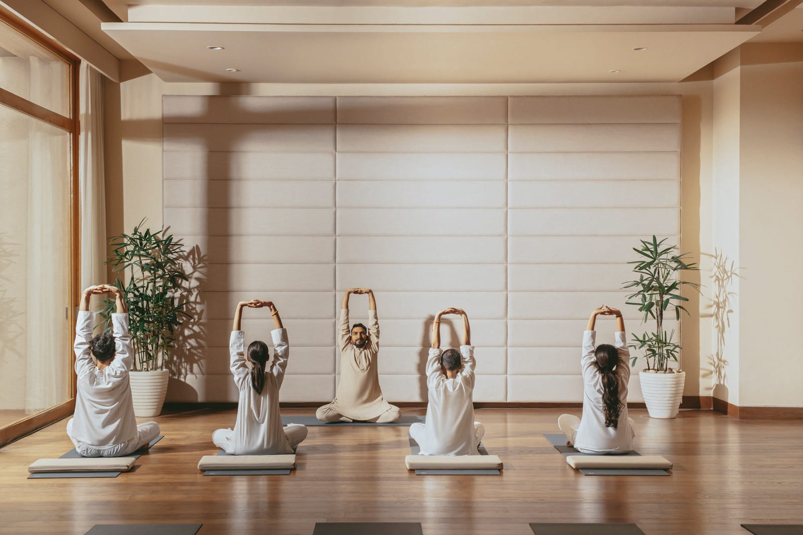 Group yoga class in bright studio, five people in white clothing performing seated side stretch with arms overhead