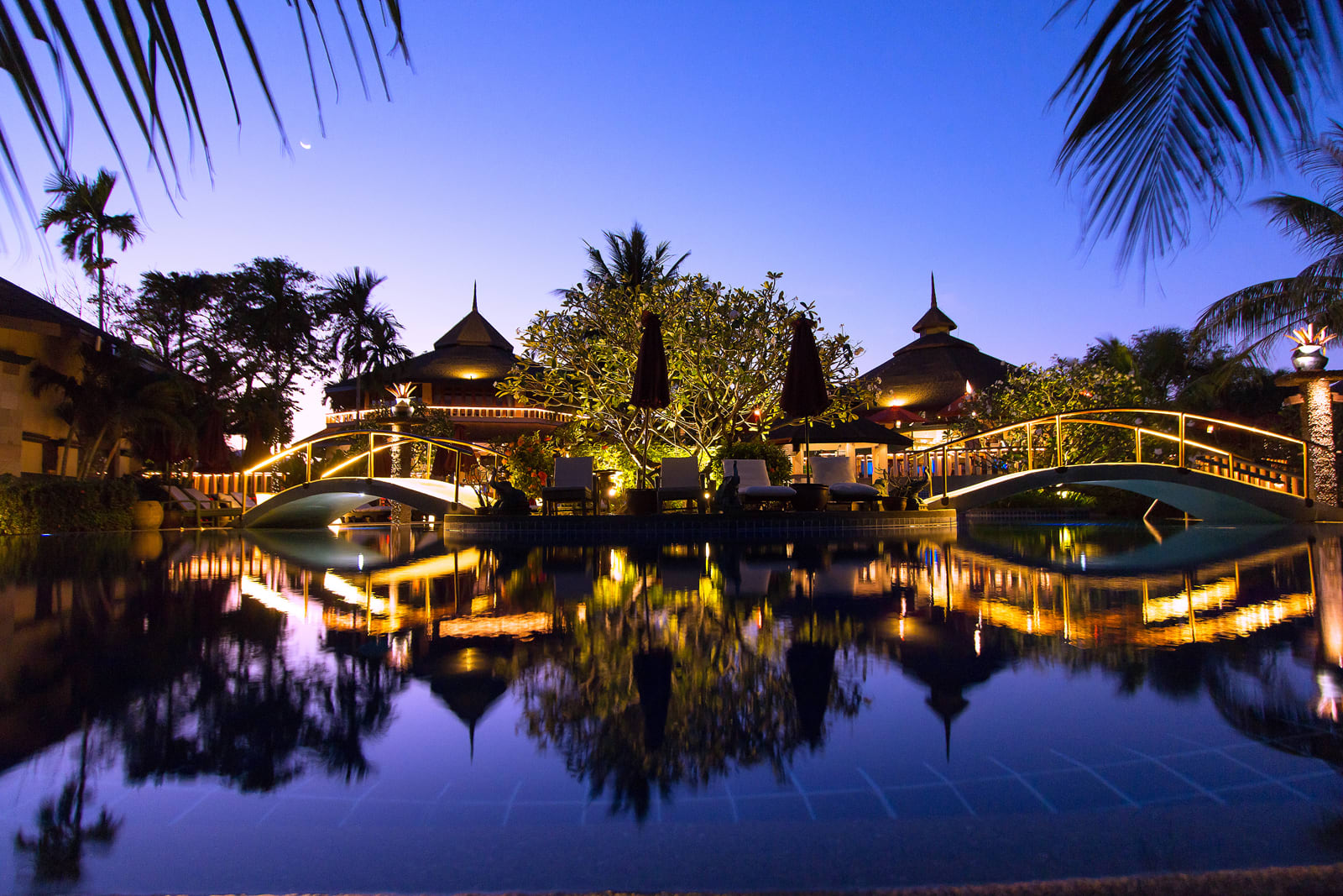 Tropical resort at twilight with illuminated pavilions, arched bridges, and calm reflecting pond surrounded by palm trees