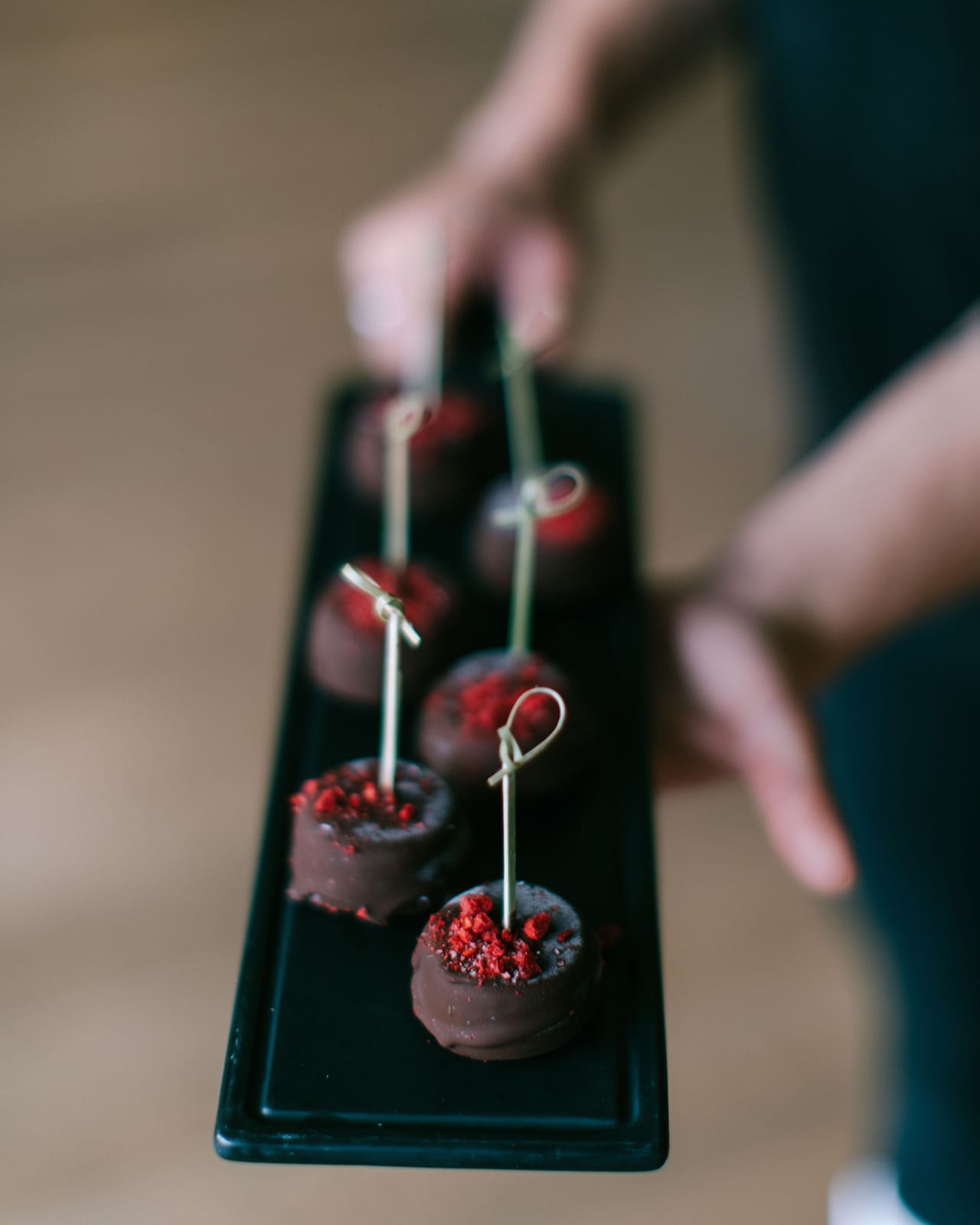 Hand holding black rectangular plate with chocolate spheres topped with red powder and decorative picks