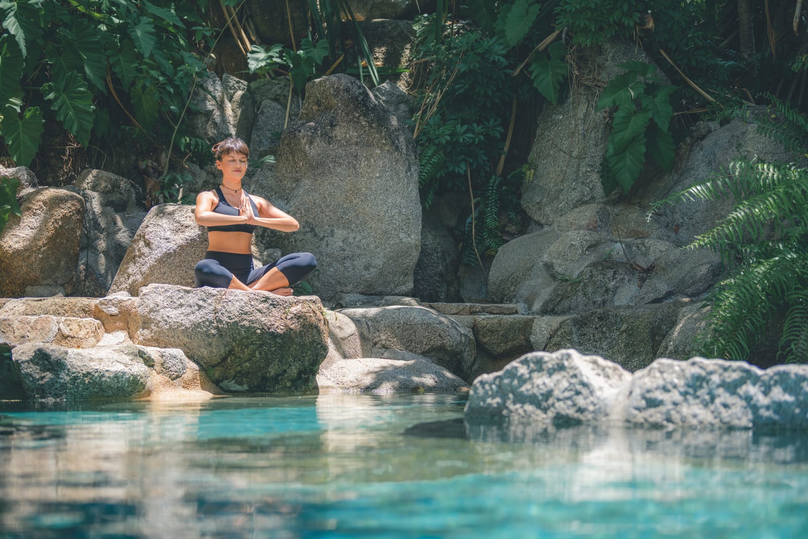 Woman in yoga pose seated on rocks beside turquoise pool surrounded by lush tropical vegetation