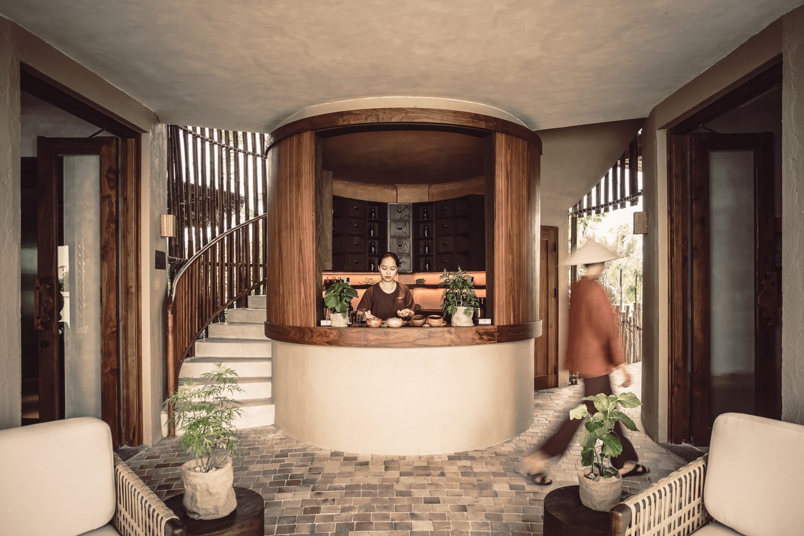 Spa receptionist at curved wooden counter with warm lighting and herringbone tile floor in wellness retreat