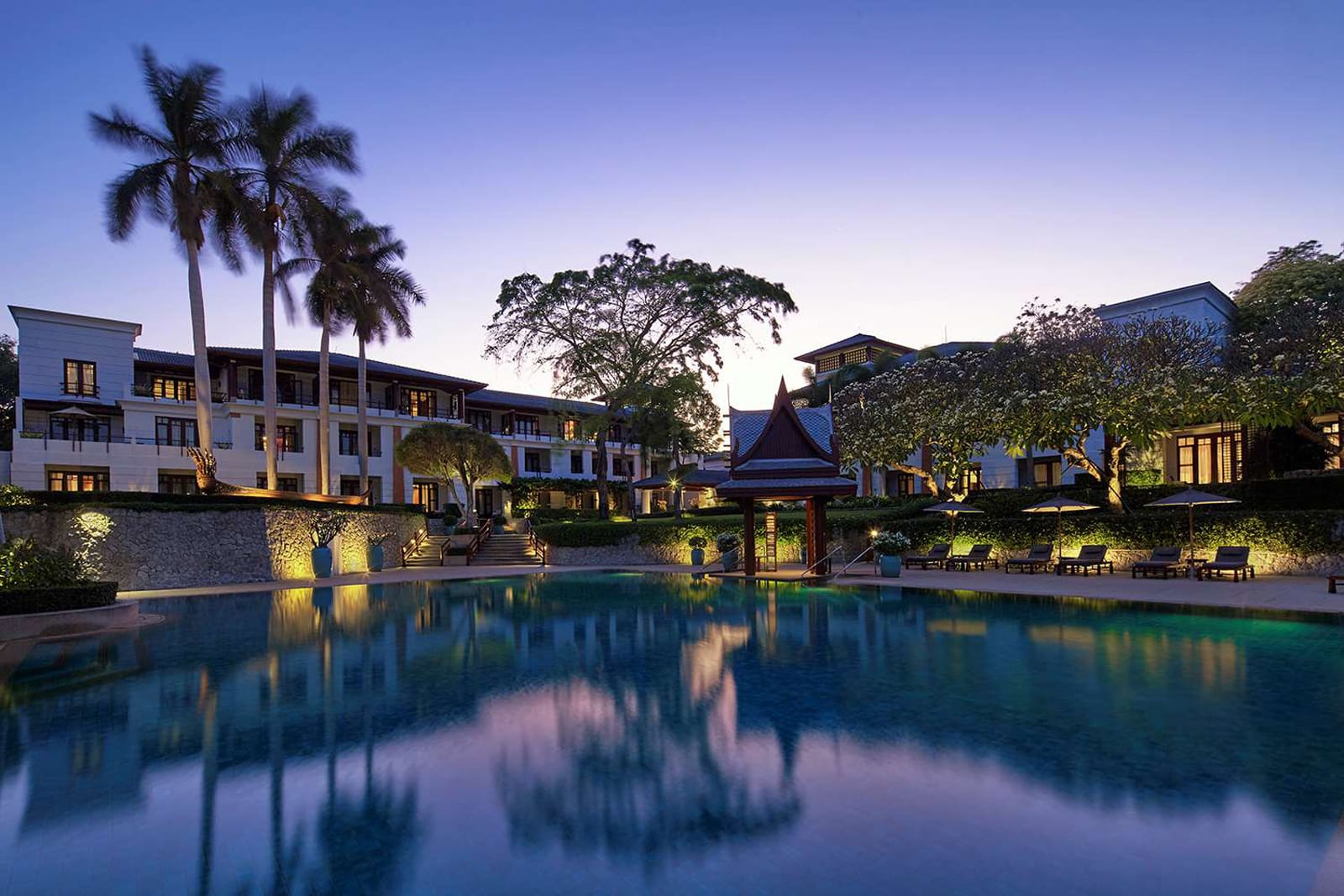 Luxury resort at twilight with illuminated white buildings, palm trees, and reflecting pool at dusk