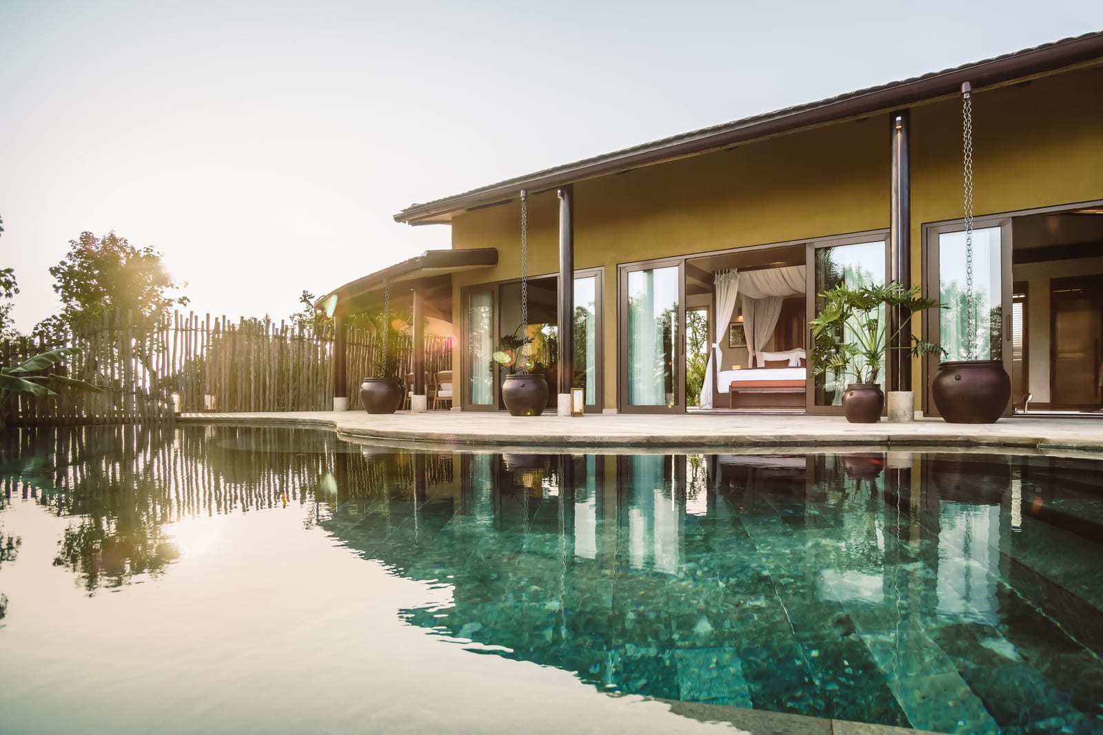 Modern luxury villa with yellow walls and large windows overlooking infinity pool at sunset