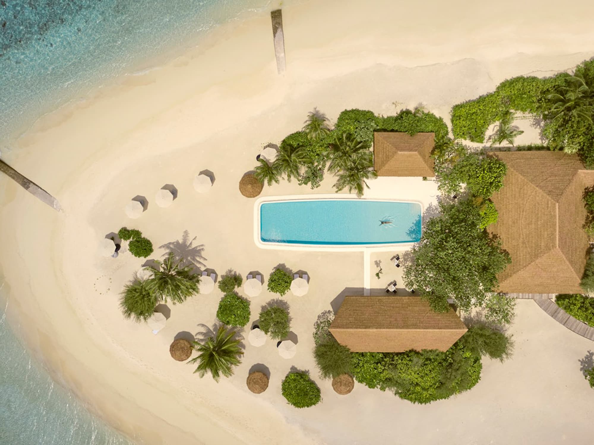 Aerial view of luxury beach resort with rectangular pool, thatched-roof buildings, and palm trees on white sand