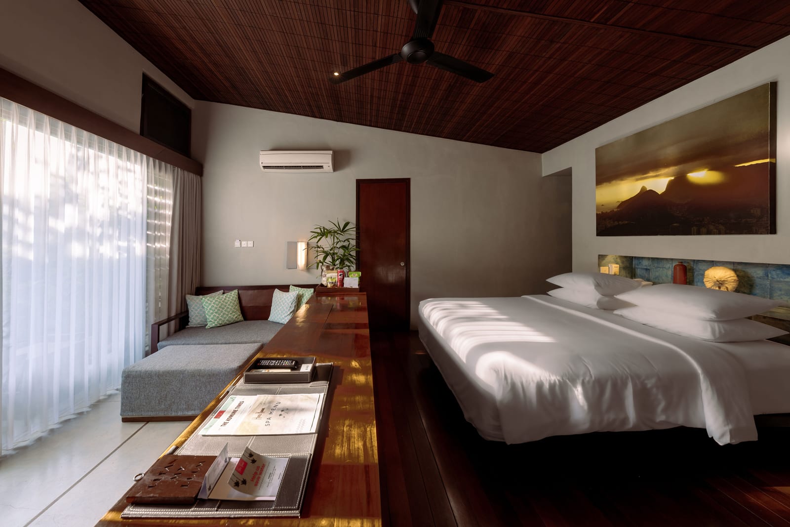 Modern hotel bedroom with wooden ceiling, queen bed, lounge area, and landscape artwork