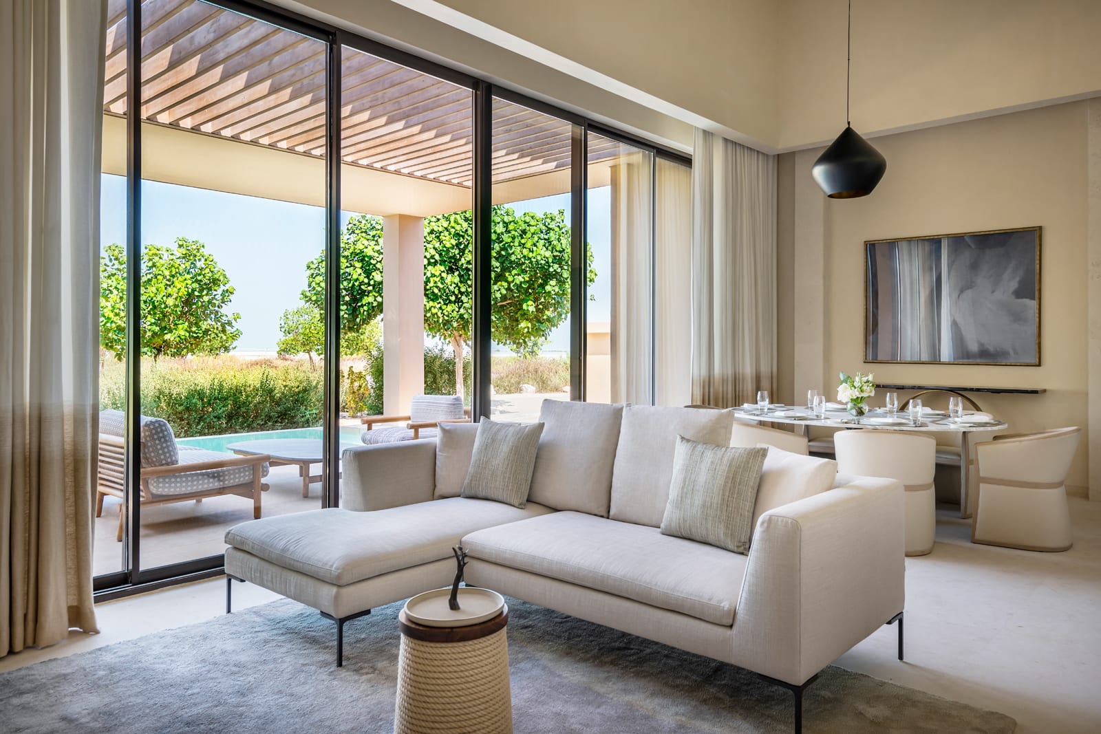 Modern living room with cream sectional sofa, black pendant light, and floor-to-ceiling windows overlooking pool and landscape
