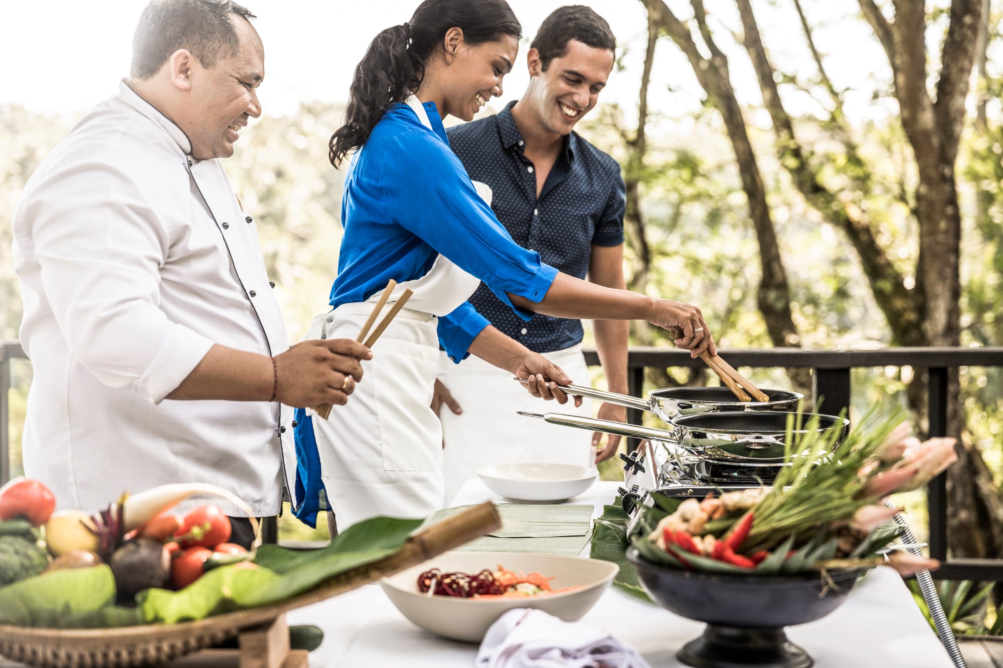 Three people cooking together outdoors with fresh vegetables and cookware on table