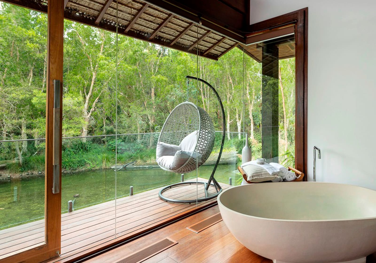 Modern bathroom with freestanding tub overlooking deck, hanging chair, and forested river view